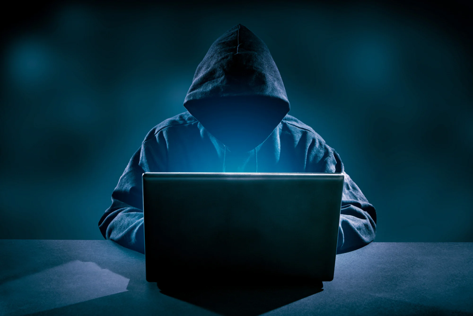 A hooded figure working at a laptop in a darkened room